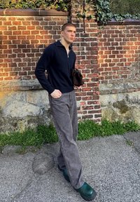 Černý pletený pulovr s polovičním zipem, šedé široké kalhoty a zelené platformové dřeváky, stojící před cihlovou zdí.