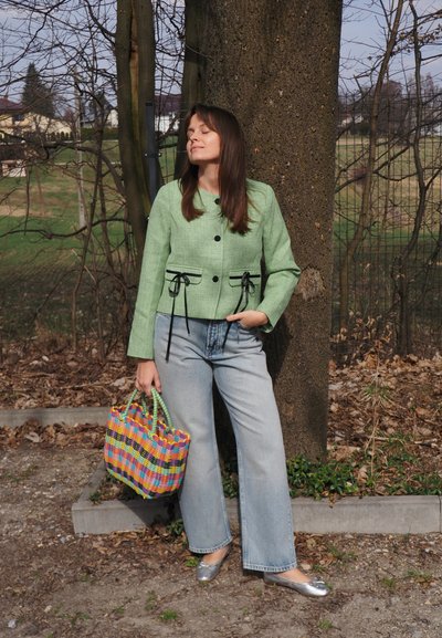 Mujer con chaqueta verde, jeans azul claro y zapatos planos plateados apoyada contra un árbol, sosteniendo una bolsa tejida de colores al aire libre.