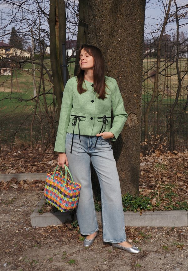 Femme en veste verte, jean bleu clair et ballerines argentées, debout contre un arbre, tenant un sac coloré tissé en plein air.