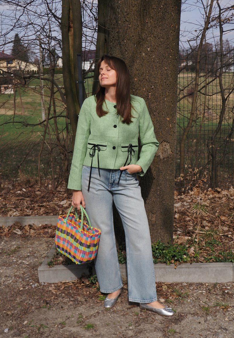 Vrouw in een groene jas, lichtblauwe spijkerbroek en zilveren ballerina's staat buiten tegen een boom, met een kleurrijke geweven tas in haar hand.