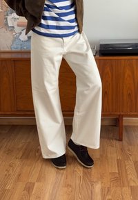 Široke bele bombažne hlače v kombinaciji s črnimi čevlji. V ozadju je lesen kabinet in modro črtasta majica.