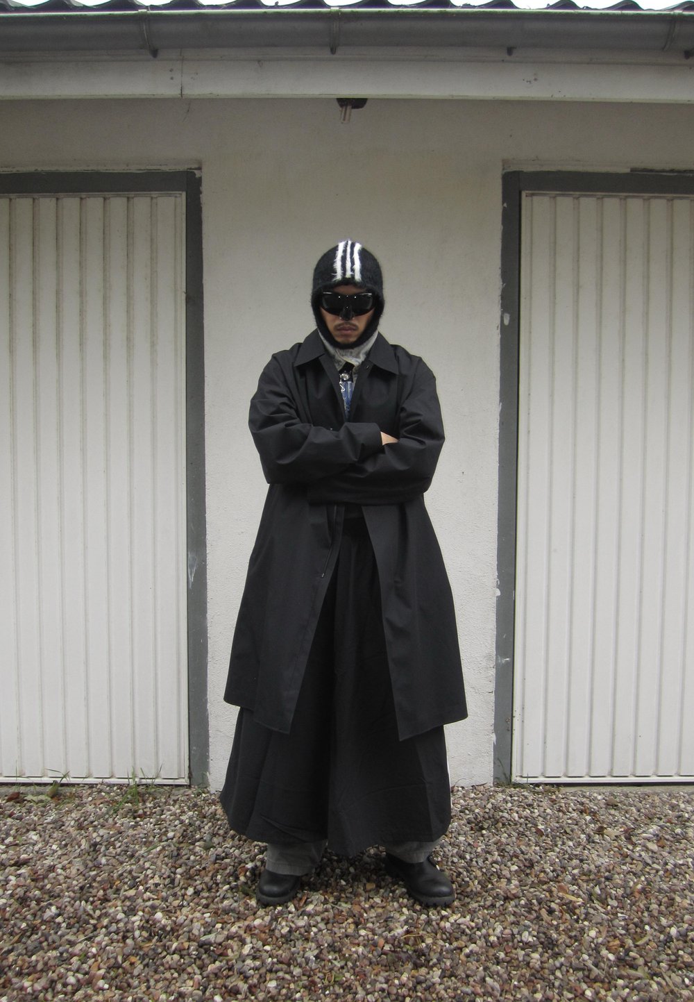 Fekete trench coat egy hosszú szoknya, szürke nadrág és fényes fekete cipők felett. Kiegészítők közé tartozik egy fekete csíkos sapka és sötét napszemüveg.
