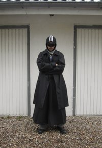 Fekete trench coat egy hosszú szoknya, szürke nadrág és fényes fekete cipők felett. Kiegészítők közé tartozik egy fekete csíkos sapka és sötét napszemüveg.