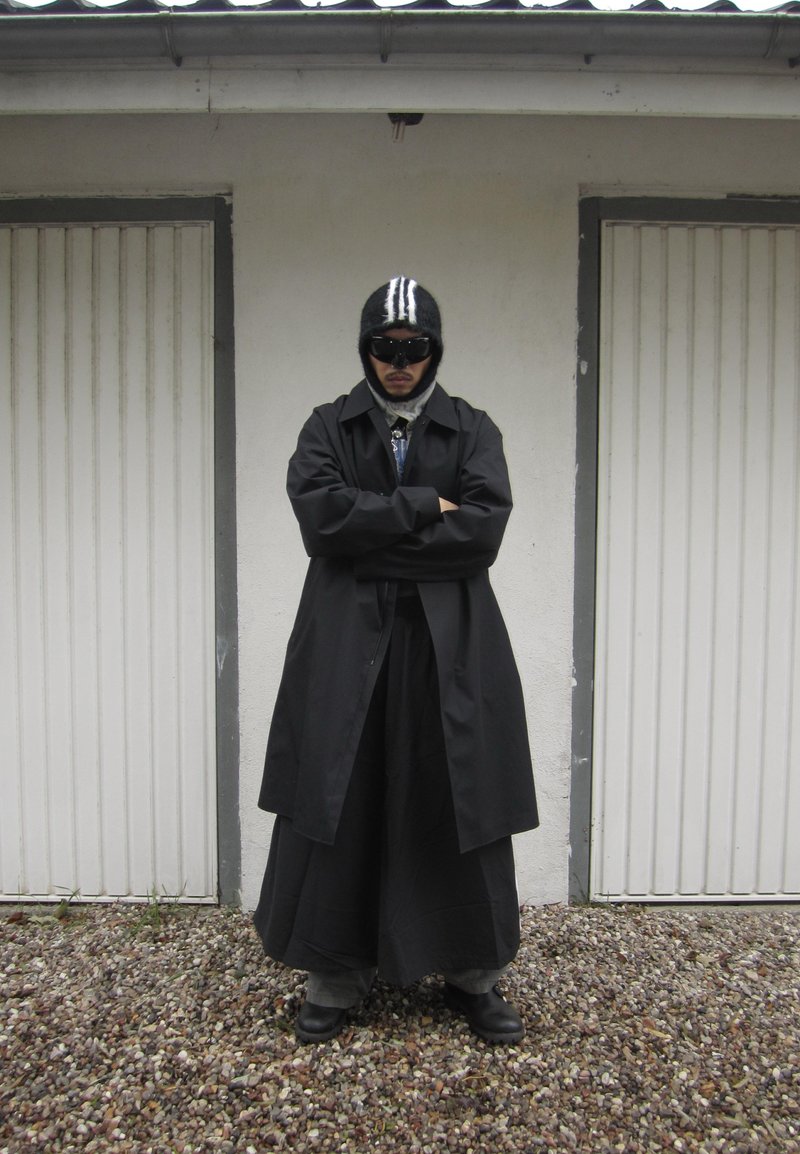 Black trench coat over a long skirt, gray pants, and shiny black shoes. Accessories include a black striped beanie and dark sunglasses.