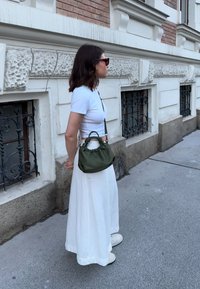 Ελιά πράσινη τσάντα ώμου με συγκεντρωμένη κορυφή, φορεμένη χιαστί. Μοντέλο με άσπρη, μακριά φούστα και κοντομάνικο μπλουζάκι, στέκεται σε πεζοδρόμιο κοντά σε τοίχο από τούβλα.