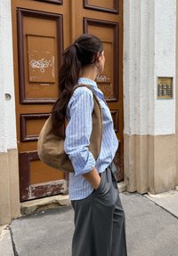 Sac en cuir suédé brun, chemise rayée bleu clair, pantalon à fines rayures grises. L'arrière-plan comprend une porte en bois brun et un mur blanc.