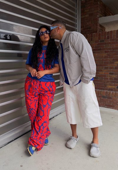 Pantalones rojos brillantes con un patrón abstracto azul, combinados con una camiseta gráfica azul. Los accesorios incluyen gafas de sol y calzado informal.