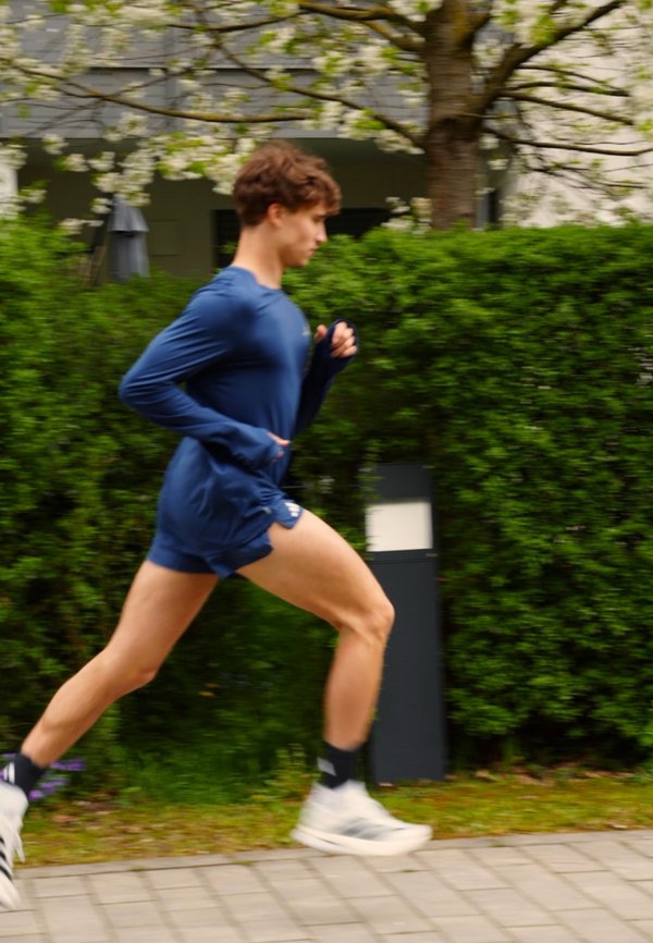 Junger Mann läuft auf einem gepflasterten Weg neben Grünflächen und einem Baum mit weißen Blüten, trägt blaue Sportkleidung und weiße Turnschuhe.