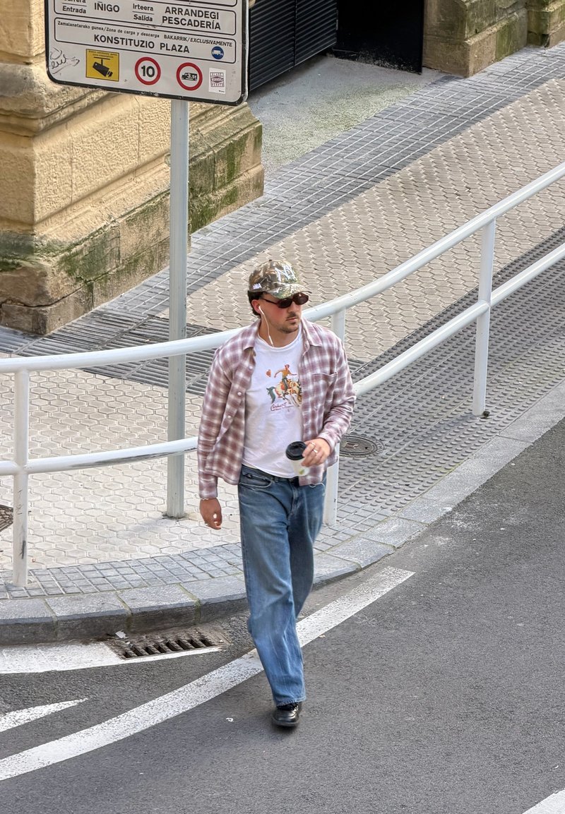 Homem vestindo camisa xadrez, jeans e boné caminhando na rua perto de uma placa de trânsito e grades metálicas brancas numa área urbana.