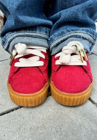 Κόκκινα πάνινα παπούτσια καπιτονέ με λευκές κορδόνες, σόλα από καουτσούκ και μεταλλικά άκρα. Φορεμένα με μπλε τζιν με γυρισμένα μπατζάκια, αναδεικνύοντας την υφή και την αντίθεση χρωμάτων.