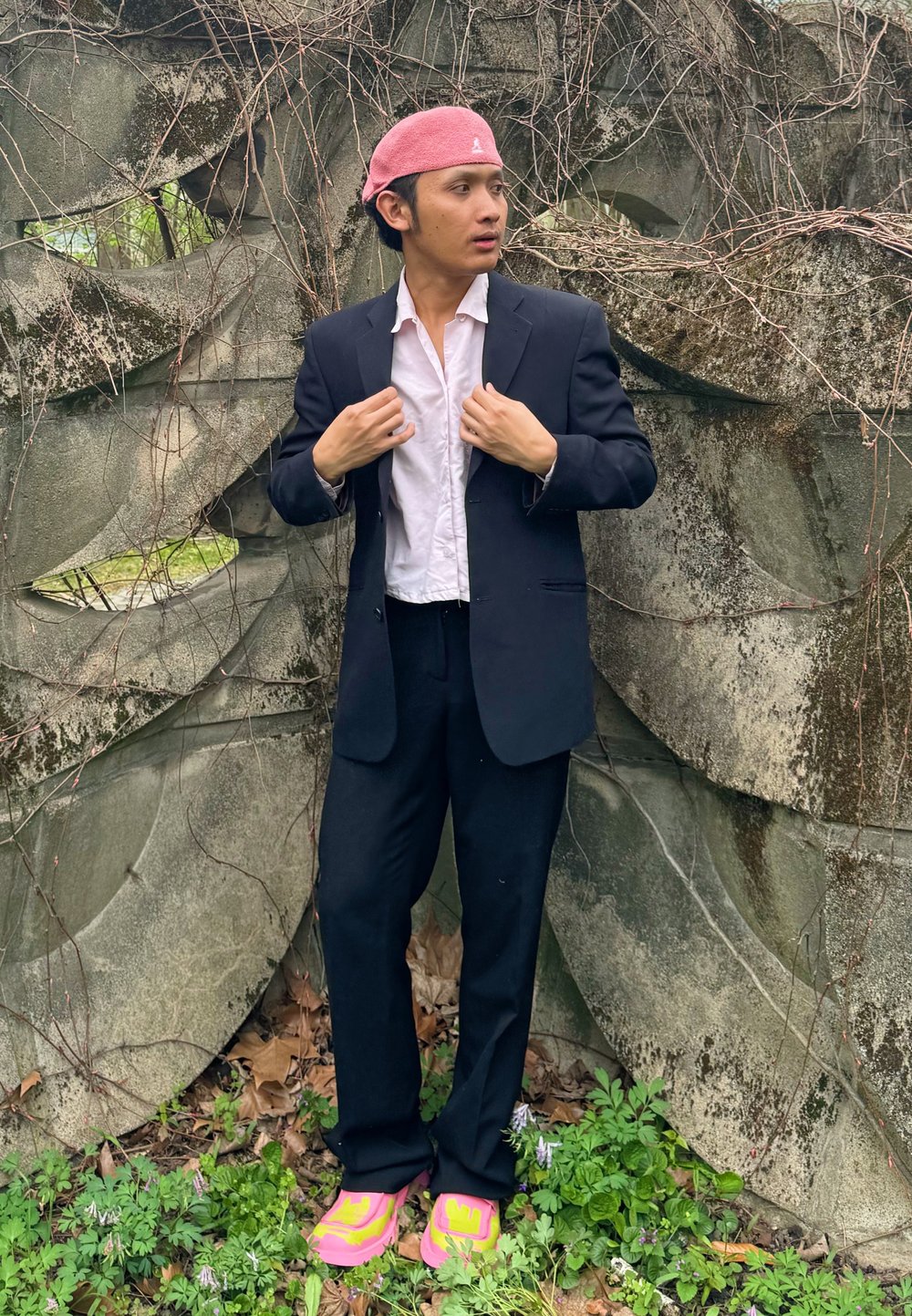Black tailored suit paired with a white button-up shirt, pink headwear, and vibrant pink trainers. Leaves and vines feature in the background.