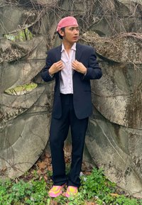 Black tailored suit paired with a white button-up shirt, pink headwear, and vibrant pink trainers. Leaves and vines feature in the background.