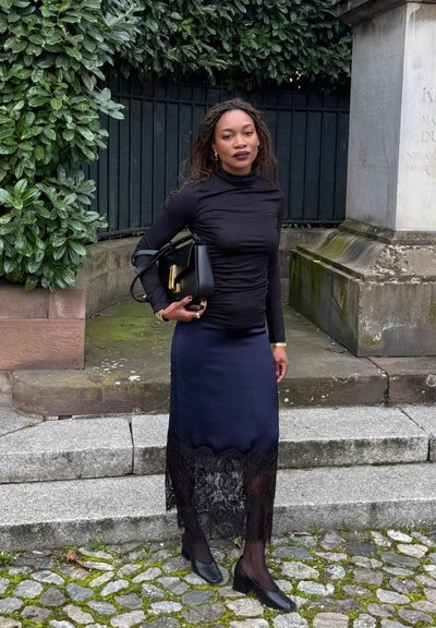 Mujer con blusa negra de manga larga y falda con encaje sostiene un bolso negro, de pie sobre escalones de piedra cerca de vegetación y un monumento.