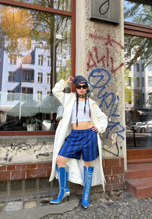 Person trägt einen weißen langen Mantel, blaue gestreifte Shorts, blaue Schnürstiefel mit hohem Absatz, schwarze Sonnenbrille und ein schwarzes gemustertes Kopftuch und steht bei Graffiti.