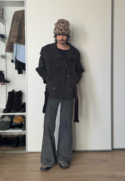 Persona con un gorro negro con estampado de leopardo, chaqueta negra de doble botonadura con tirantes y jeans grises de pierna ancha, de pie en una habitación con estanterías y zapatos.