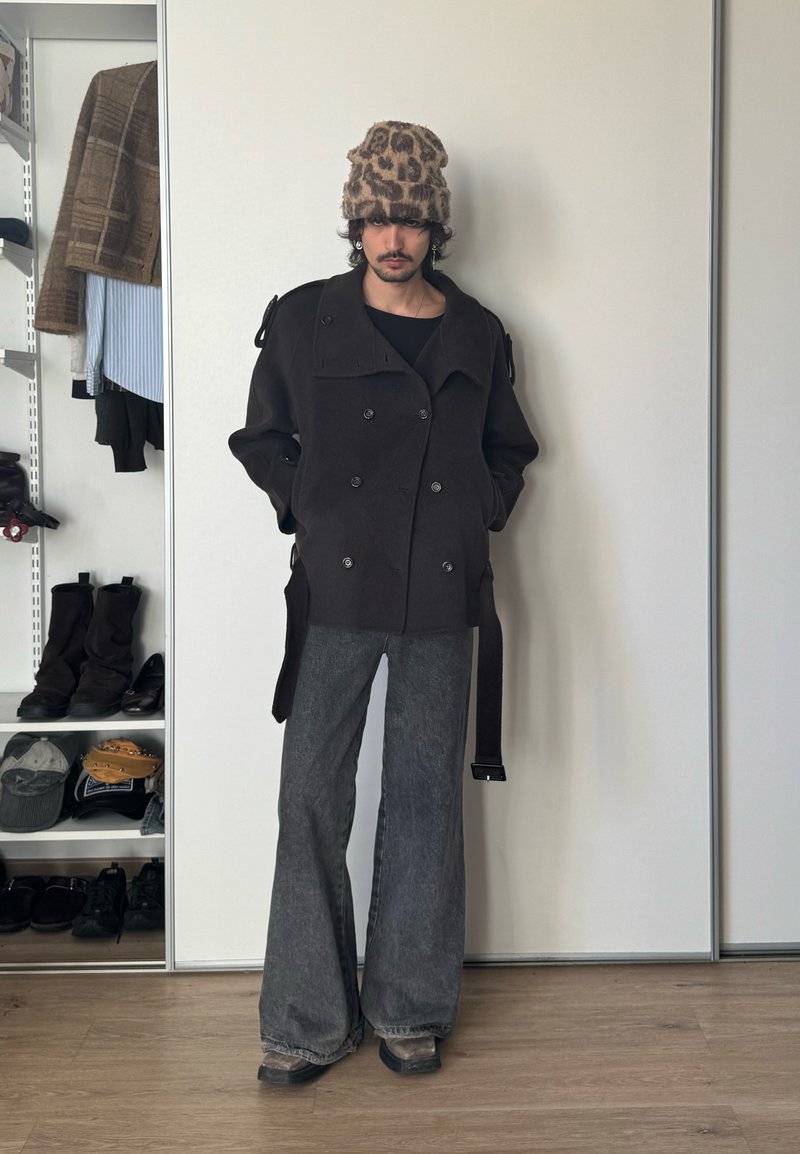 Inimene, kes kannab leopardimustrilise beanie't, musta kahekarvalist rihmadega jopet ja laiade säärega halli teksapükse, seisab toas, kus on riiulid ja jalanõud.