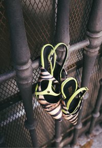 Chaussures à lanières slingback rayées noir et marron avec une lanière accentuée vert néon, suspendues à une rampe métallique grise devant un fond en maille.