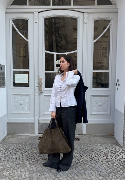 Mujer con blusa blanca y pantalones oscuros sosteniendo un gran bolso marrón y una chaqueta azul marino, de pie sobre adoquines frente a una puerta doble blanca y ornamentada.
