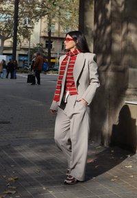 Lichtgrijs op maat gemaakt pak met wijde broekspijpen, gecombineerd met een rood en beige gestreepte sjaal. Felrode bril en casual schoeisel.