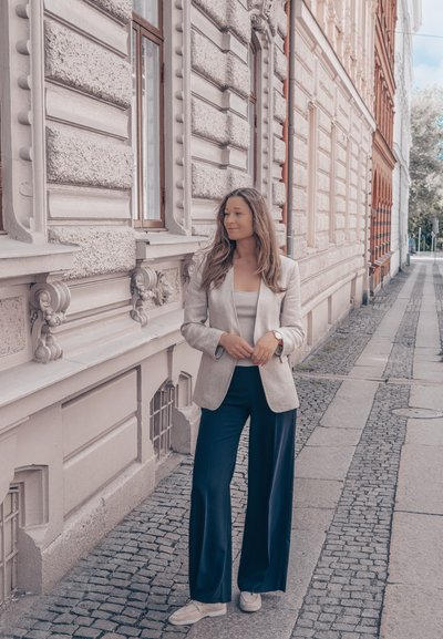 Un blazer gris claro sobre una blusa blanca, combinado con pantalones anchos de un azul marino oscuro y zapatos beige, de pie junto a detalles arquitectónicos ornamentados.