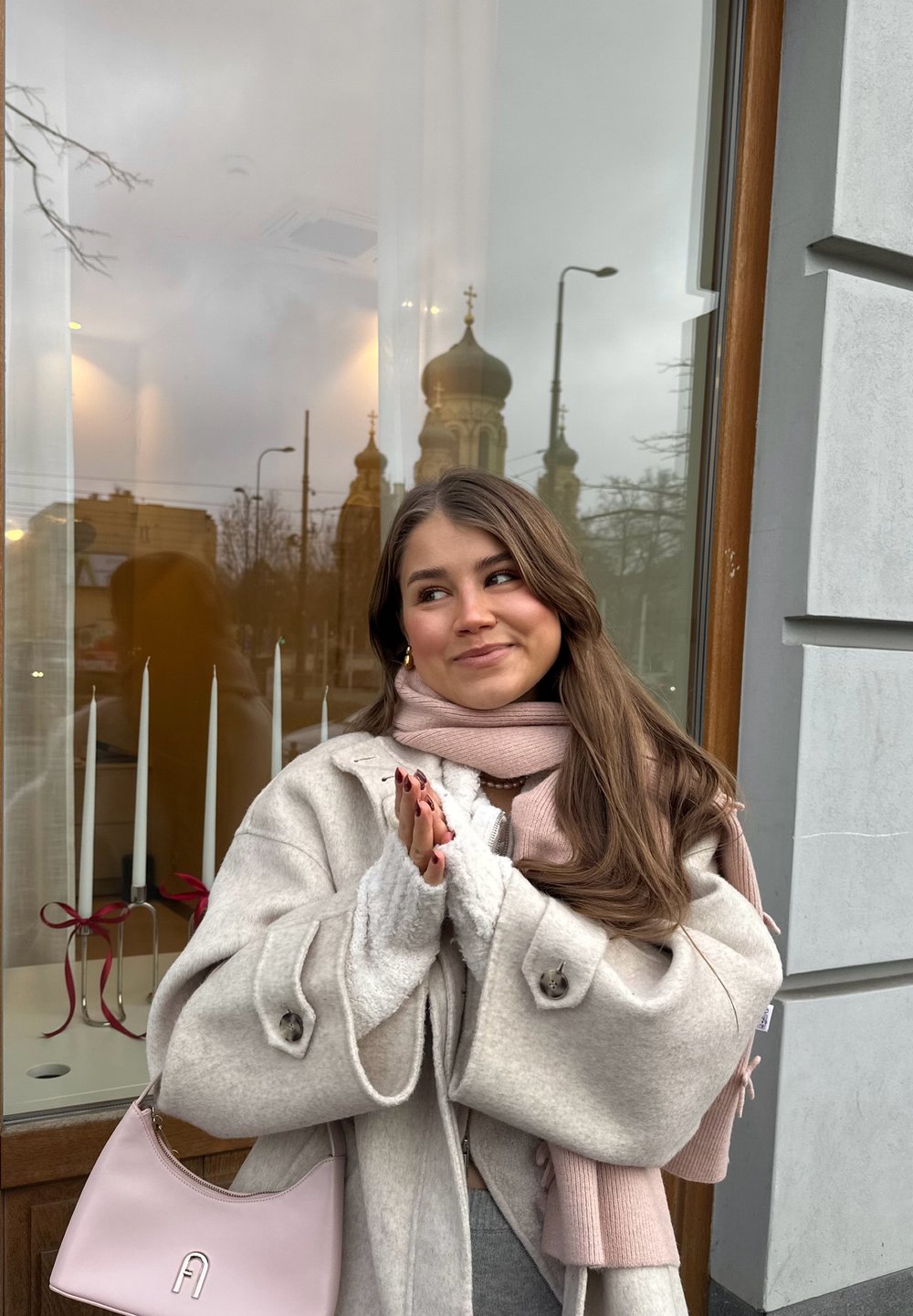 Jeune femme en manteau beige et écharpe rose applaudissant, debout devant un bâtiment avec une fenêtre reflétant des dômes d'église et des lampadaires.