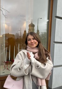 Jeune femme en manteau beige et écharpe rose applaudissant, debout devant un bâtiment avec une fenêtre reflétant des dômes d'église et des lampadaires.