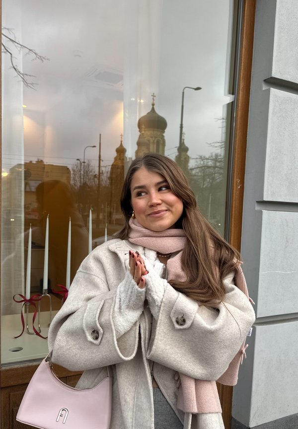 Jeune femme en manteau beige et écharpe rose applaudissant, debout devant un bâtiment avec une fenêtre reflétant des dômes d'église et des lampadaires.