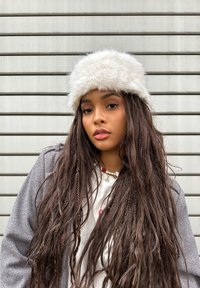 Λευκό καπέλο από ψεύτικη γούνα με σφαιρικό σχέδιο, φορεμένο πάνω από μακριές, σκούρες πλεξίδες. Το ντύσιμο περιλαμβάνει ένα γκρίζο σχέδιο μπουφάν και στρωτές αλυσίδες.