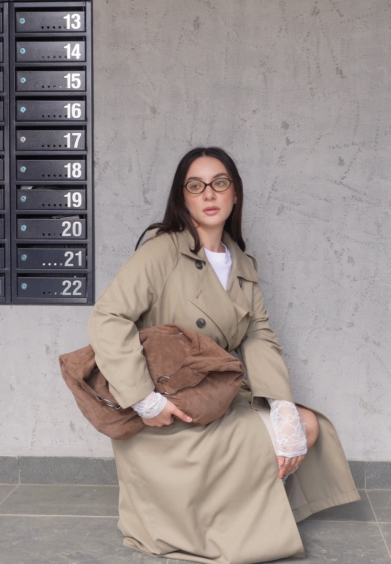 Femme portant des lunettes et un trench-coat beige, tenant un grand sac marron, assise près de boîtes aux lettres numérotées noires sur un mur gris.