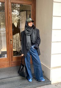 Overdimensioneret sort blazer, grå tørklæde, afslappet sort top, vide blå jeans og blå sneakers. Sort tote bag. Butiksfacade i baggrunden.