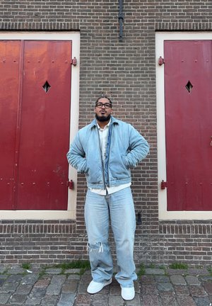 Hombre con gafas que lleva chaqueta y pantalones de mezclilla azul claro, de pie con las manos en los bolsillos frente a una pared de ladrillos con dos puertas rojas.