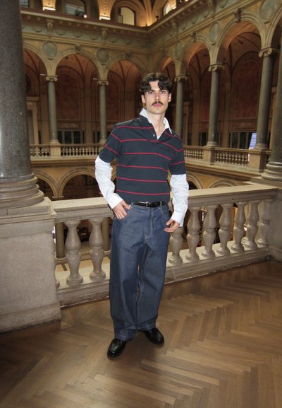 El modelo lleva una camiseta de polo a rayas oscuras con un cuello blanco, combinada con unos jeans de denim azul sueltos y zapatos negros, de pie cerca de un balcón ornamentado.