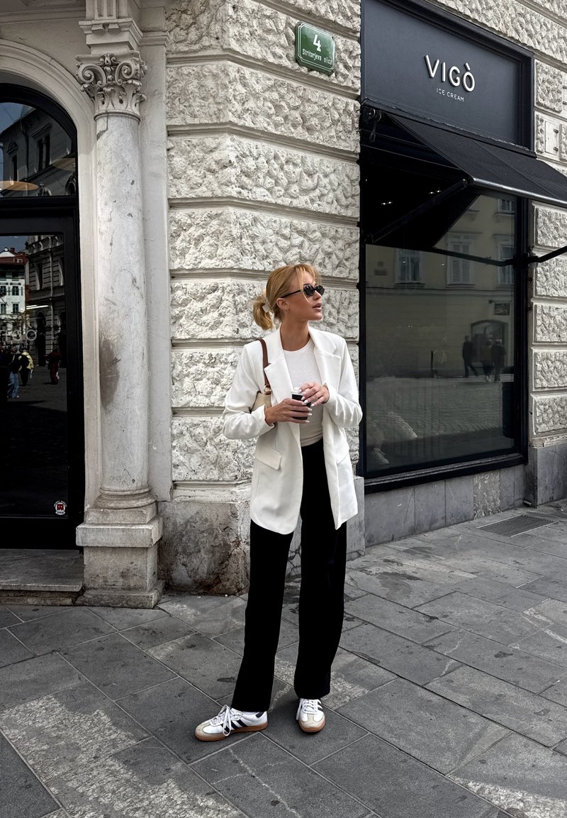 Femme en blazer blanc, pantalon noir et baskets, tenant une tasse de café, debout près d'un bâtiment en pierre avec une enseigne de la boutique de glaces Vigò.