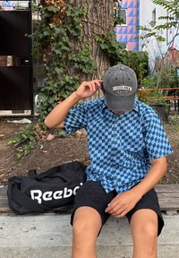 Osoba u plavoj košulji s kariranim uzorkom i sivoj Brooklyn kapi sjedi na drvenoj klupi, prilagođavajući kapu, uz crnu Reebok torbu pored sebe.