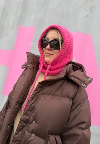 Brūna pūkaina jaka ar metālisku aparatūru, rozā adīts kapuce ar auklām un lieli melni saulesbrilles pret pelēku fonu.