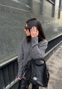 Szürke kötött pulóver bordázott mandzsettákkal, fekete bőr kézitáska fémes díszítésekkel és texturált felületek a háttérben.