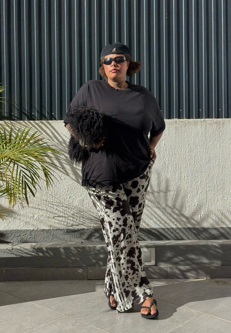 Pessoa vestindo um boné preto, óculos de sol, camiseta preta, calças com padrão de vaca e sandálias pretas, segurando um cachorro preto e peludo, em pé ao ar livre.