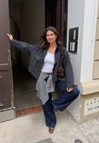 Črna karirana jakna, bela majica brez rokavov, siva karirana obleka in razširjene modre jeans hlače. Rjavi usnjeni čevlji in torba na čez ramo. Ozadje odprtih vrat.