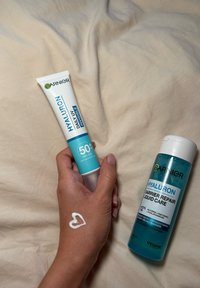 Garnier Hyaluron Daily UV usynlig kremtube vises i hånden med en blå barriere-reparerende væskeflaske ved siden av. Huden viser en hjerteformet krem.