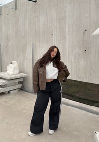 Brązowa skórzana kurtka, szara krótka bluza z kapturem, czarne spodnie dresowe z białymi paskami oraz białe trampki. Model pozujący na tle betonowej ściany.