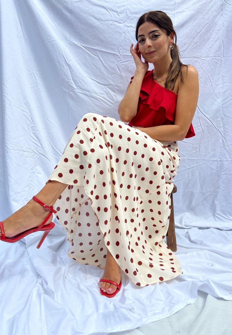 Piros, egy vállon viselhető felső fodros részletekkel, hosszú krémszínű szoknyával párosítva, amelyen piros pöttyök találhatók. Piros magassarkú szandálokkal kiegészítve.