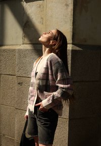 Sweter narzutowy w miękkich, pastelowych kolorach w kratę, z białymi akcentami i guzikami, zestawiony z ciemnoniebieskimi szortami dżinsowymi i czarną torbą.