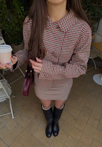 Robe-chemise à carreaux vichy rouge et blanc avec un large col, associée à des bottes noires montant jusqu'aux genoux. Tenant une tasse de café et un sac bordeaux.