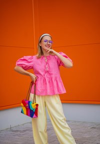 Różowa bluzka peplum z bufiastymi rękawami i wiązaniem z przodu, zestawiona z jasnymi żółtymi spodniami o szerokich nogawkach i wielokolorową torebką.