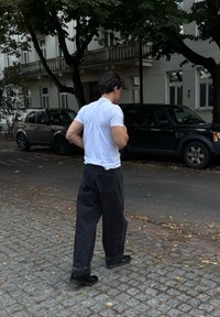 Uomo in t-shirt bianca e pantaloni neri larghi che cammina su un marciapiede in ciottoli vicino a SUV parcheggiati e edifici residenziali con alberi sopra.