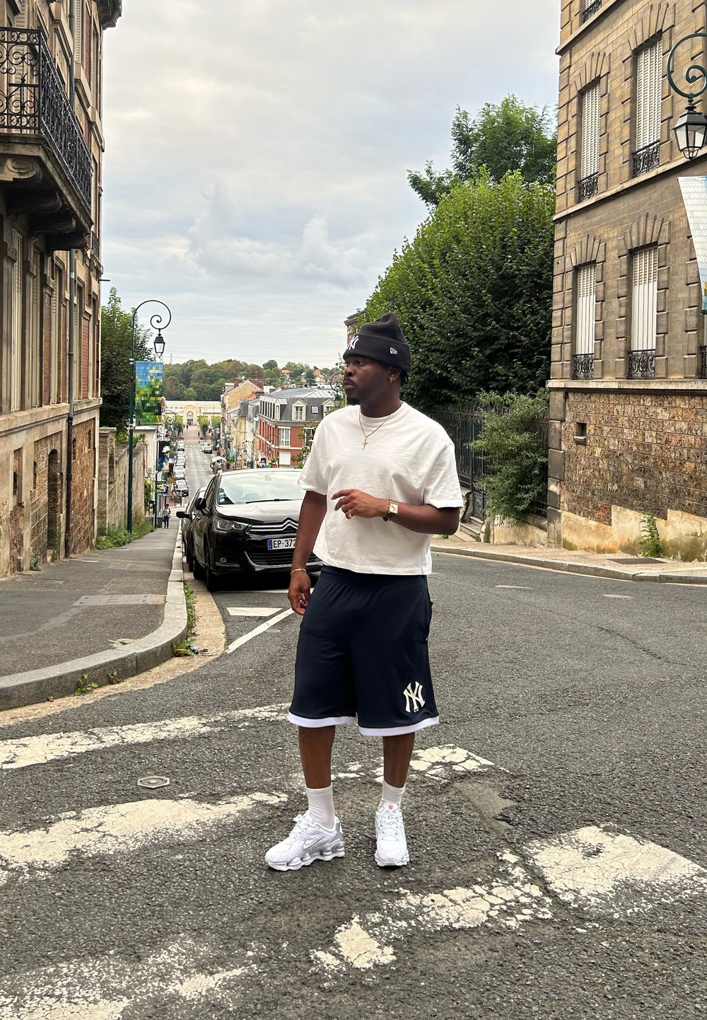 Λευκό κοντομάνικο πουκάμισο, ναυτικές σορτς με λευκό περίγραμμα και λευκά sneakers. Αστική σκηνή με κτίρια και ένα αυτοκίνητο στο παρασκήνιο.