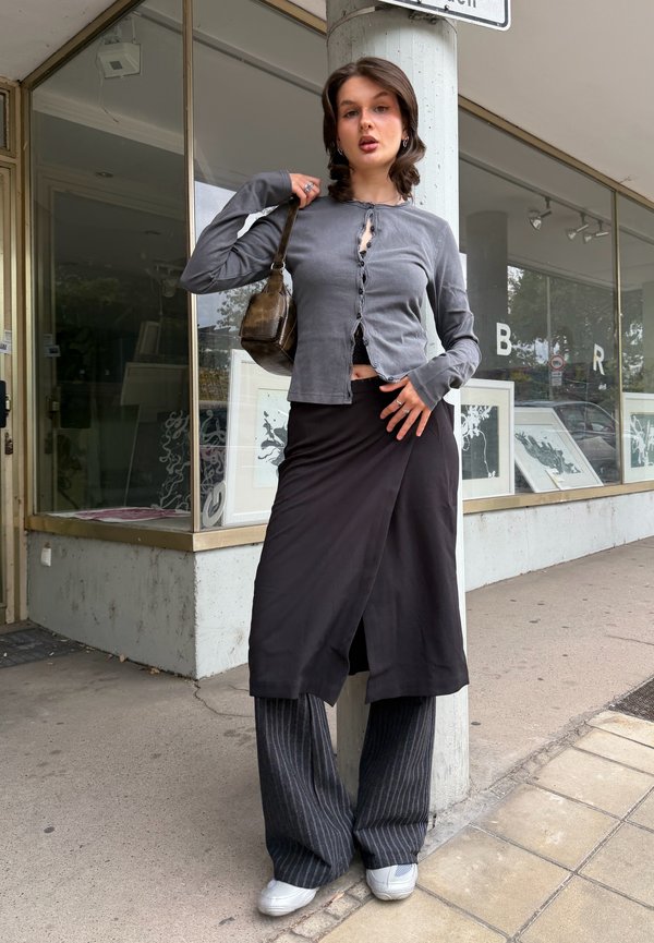 Mujer joven con falda larga negra sobre pantalones de rayas y blusa gris con botones, sosteniendo bolso marrón de hombro, de pie en la acera de la ciudad.