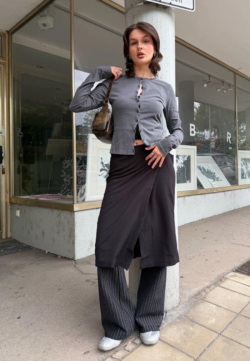 Mujer joven con falda larga negra sobre pantalones de rayas y blusa gris con botones, sosteniendo bolso marrón de hombro, de pie en la acera de la ciudad.