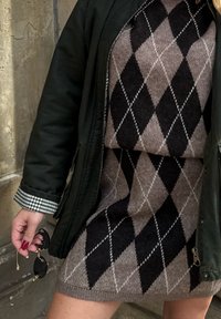 Strickkleid mit braun-schwarz kariertem Muster, kombiniert mit einer grünen Jacke. Ein schwarzer Sonnenbrillenhalter in der Hand fügt ein Detail hinzu.