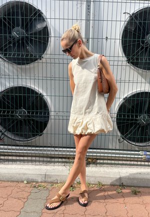 Vestido sin mangas de color crema con un dobladillo con volantes. Combinado con sandalias marrones y un pequeño bolso marrón. El fondo presenta ventiladores industriales.